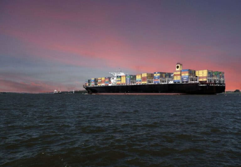 A large cargo ship carrying multicolored shipping containers, sailing on the ocean with 
a beautiful sunset sky in the background.